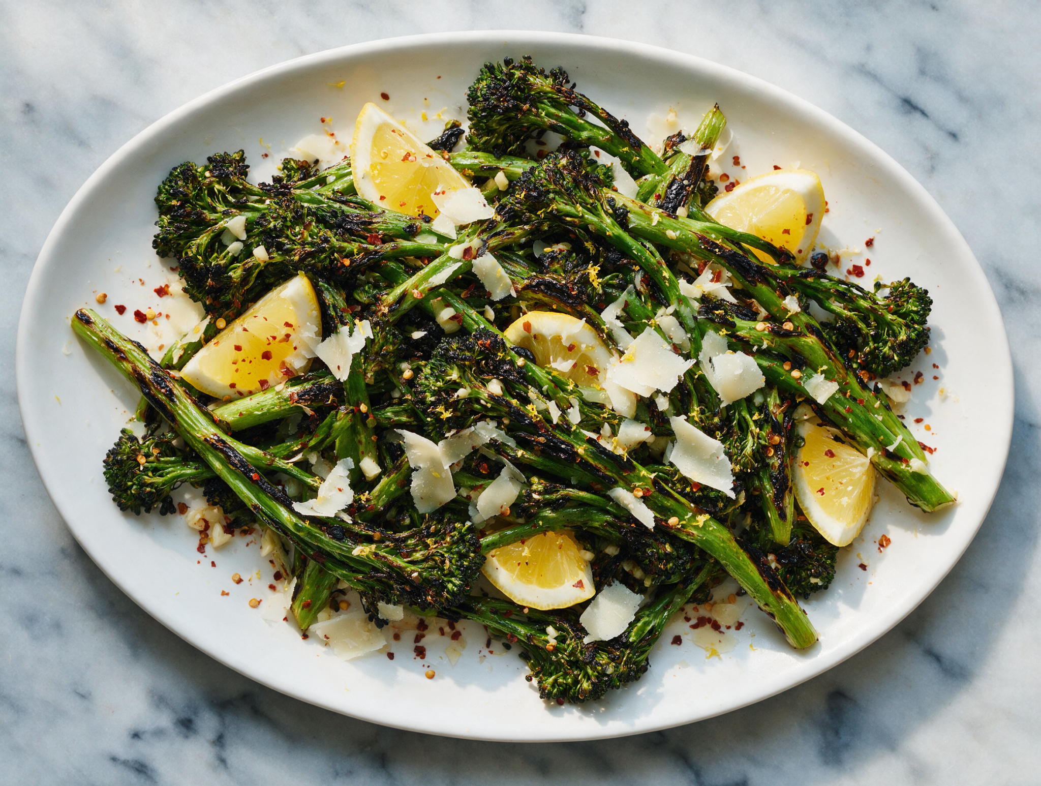 Overhead shot of charred broccolini piled on a white platter with lemon wedges, chili flakes, and shaved parmesan under soft window light.