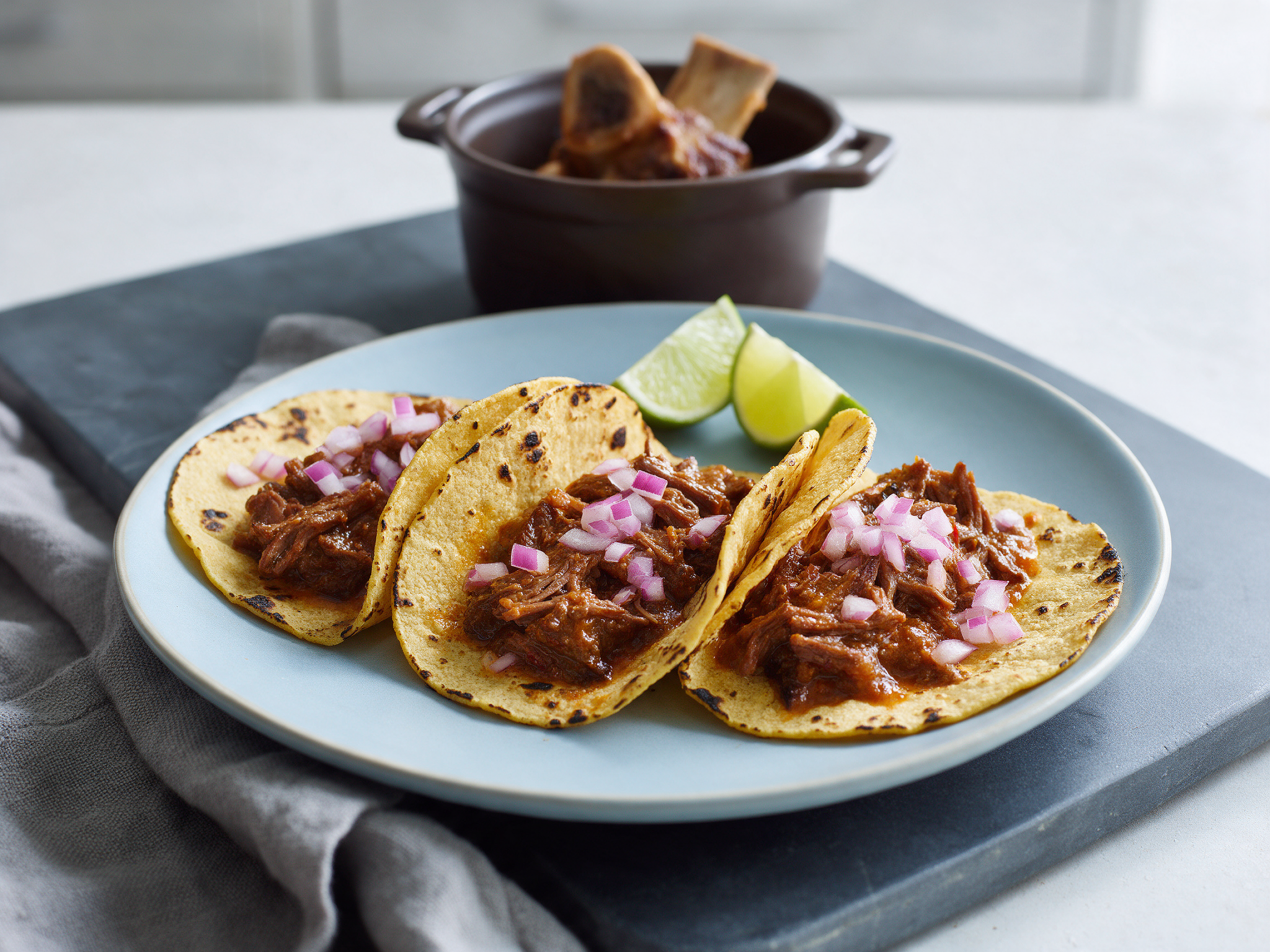 Charred corn tortilla tacos filled with shredded short rib, raw onion and lime wedges on a slate board